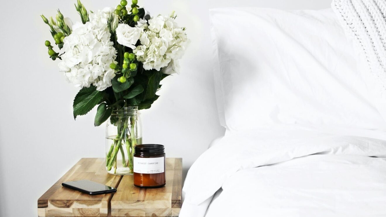 Natural wood nightstand with fresh flowers, candle, and phone next to bed.