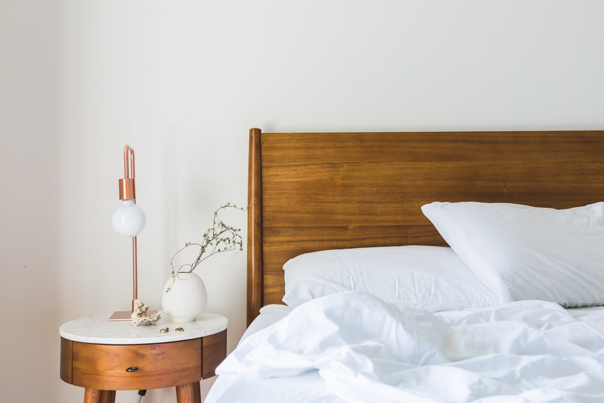 Bed with white bedding and wood headboard next to white and wood nightstand with lamp and vase.