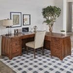 A home office with a brown hardwood desk with curved sides, a white upholstered chair, a plaid rug, industrial decor, and 2 plants.