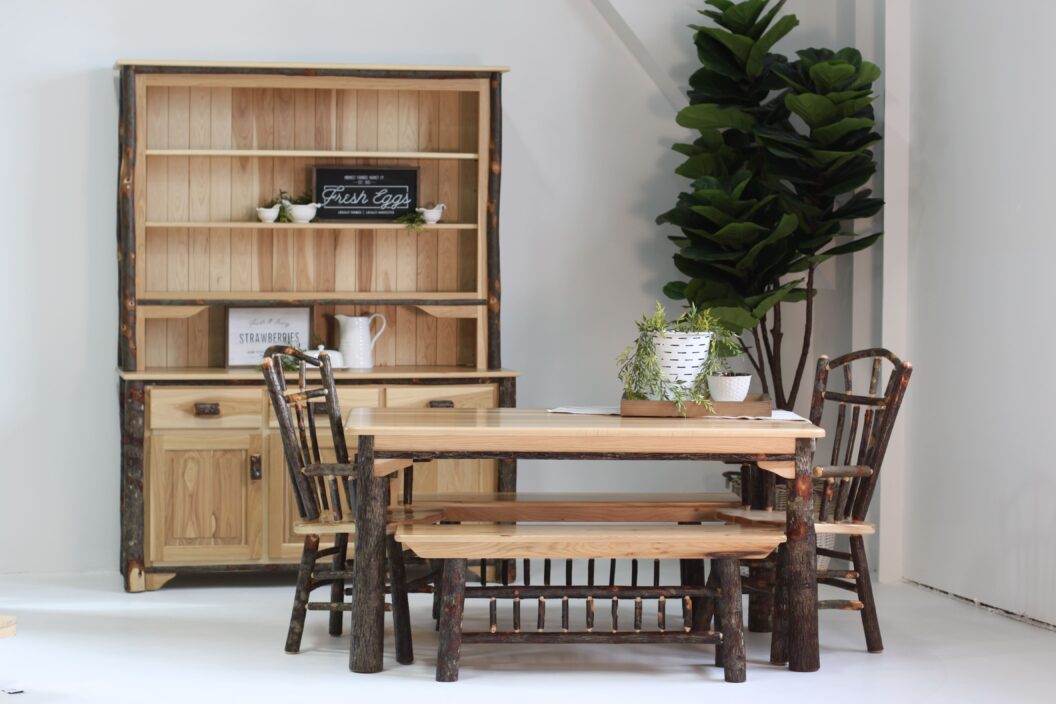 Lodge dining table with 2 benches and 2 arm chairs, lodge open shelf hutch, and a large plant in the right corner.