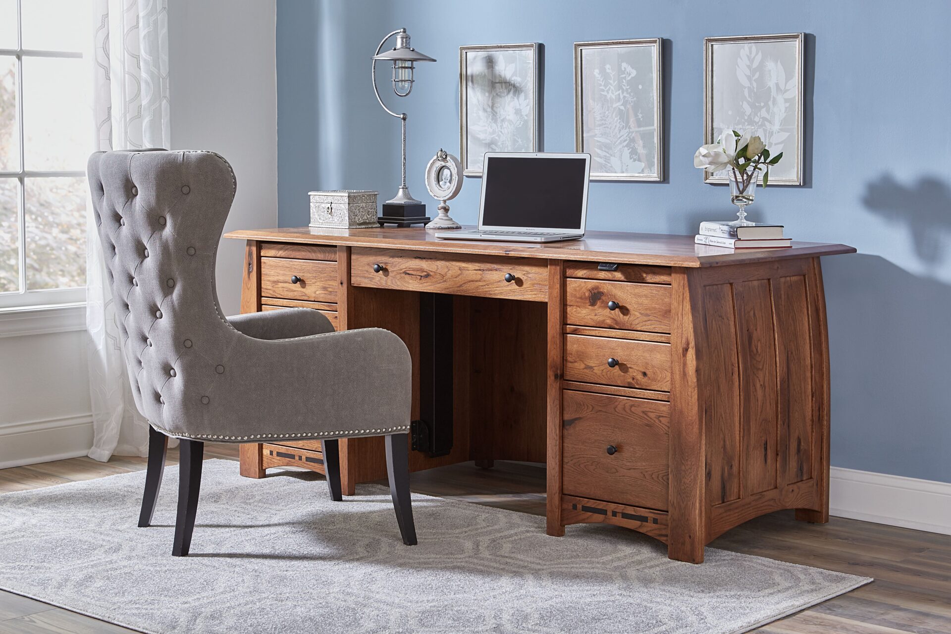 Wood desk with curved sides in a room with blue walls, white trim, brown plank flooring, a gray office chair, and a large gray and white rug.