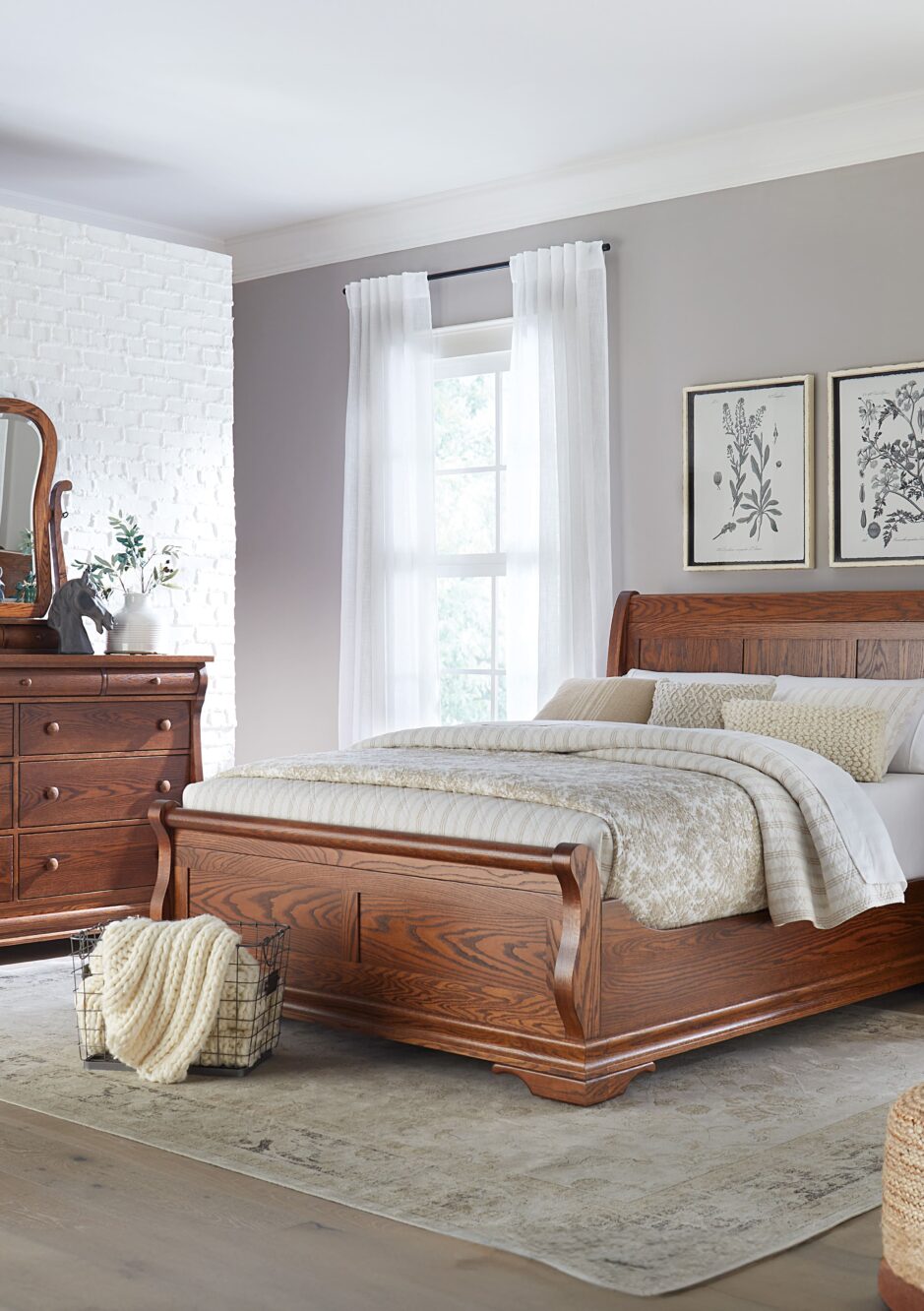 Bedroom with brown hardwood sleigh bed frame with white bedding, a brown hardwood long 6-drawer dresser with a curved mirror, light gray walls, light hardwood flooring with a large white rug, and windows with white sheer curtains.