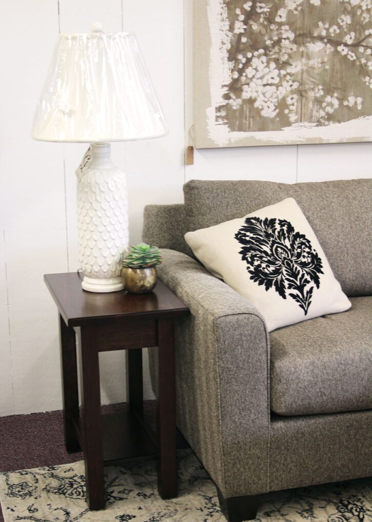 Close up of a light gray couch with a white and black pillow and sitting next to a dark brown end table with a large white lamp.