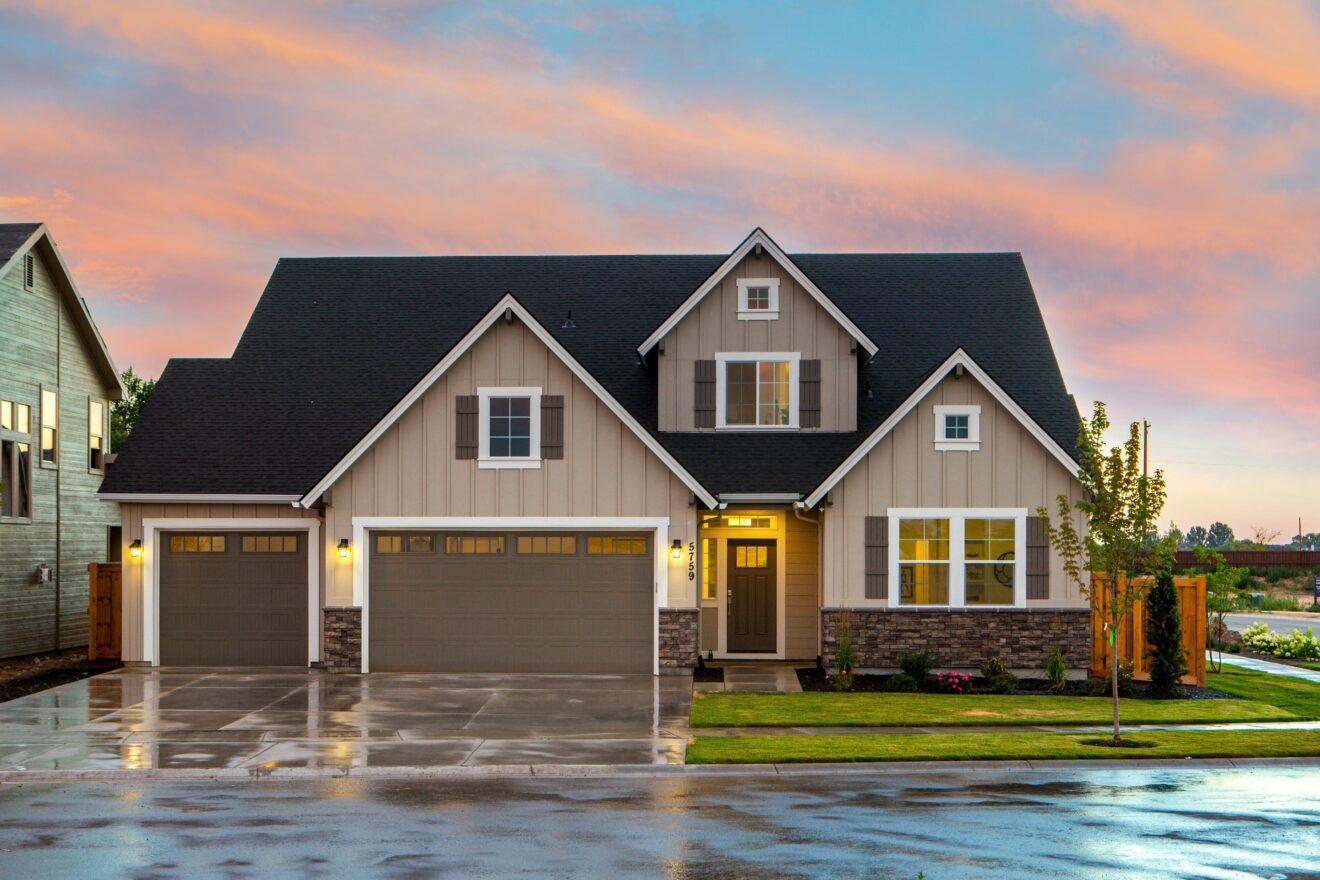 Front exterior of a house with 2 garages, stone accents, black roofing, brown shutters, white trim, and tan siding.