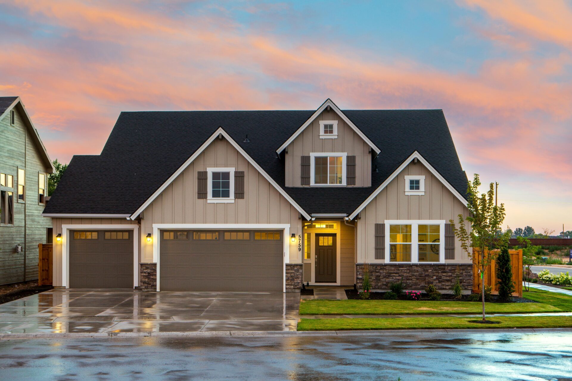 Front exterior of a house with 2 garages, stone accents, black roofing, brown shutters, white trim, and tan siding.